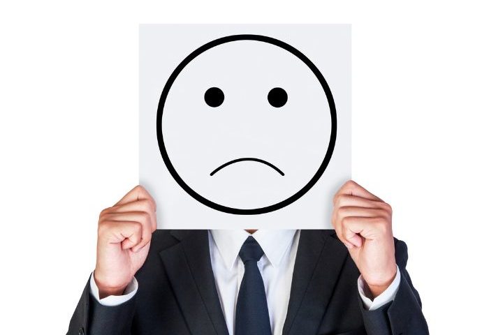 A person in a suit holding a white square with a sad face drawn on it in front of their face.